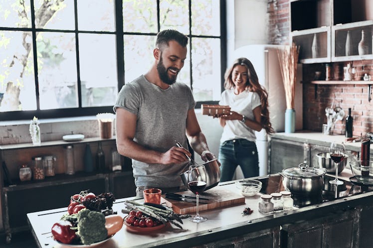 A young, happy couple enjoys wine and prepares a meal in their sunny kitchen.