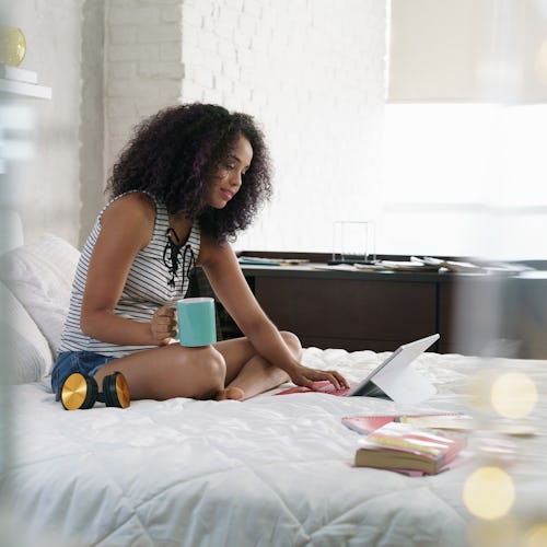 African american college student doing homework in bed at home, young black woman preparing school t...