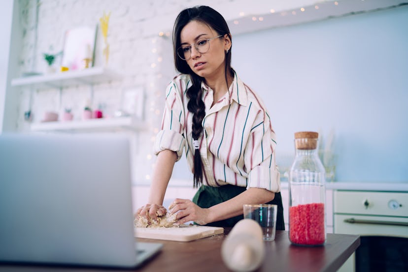 Charming black haired woman in striped shirt and glasses kneading dough while watching food recipe t...