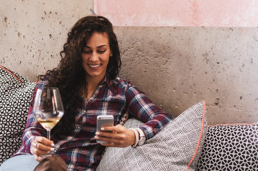 Beautiful young woman text messaging with her smart phone and holding glass of wine, sitting on sofa...