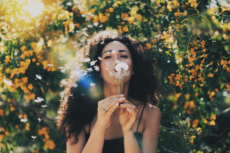 A woman blows out a dandelion in front of orange flower bushes on a sunny day.