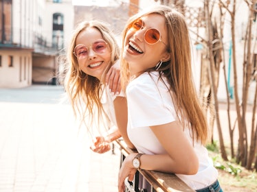 Two young women in white T-shirts and colorful sunglasses smile outside on a sunny day.