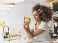 A woman in a white T-shirt smiles at her kitchen table while holding up a yellow Easter egg she pain...