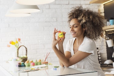 A woman in a white T-shirt smiles at her kitchen table while holding up a yellow Easter egg she pain...