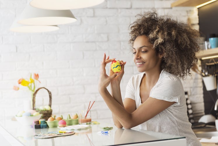A woman in a white T-shirt smiles at her kitchen table while holding up a yellow Easter egg she pain...