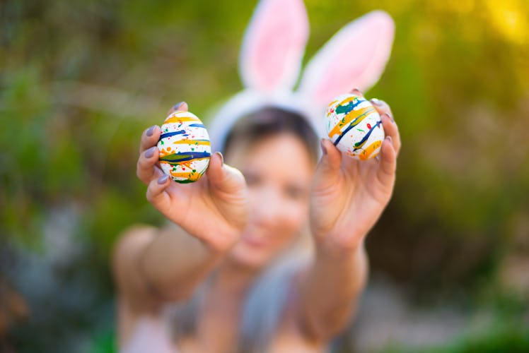 Young woman wearing bunny ears holds up two decorated Easter eggs.