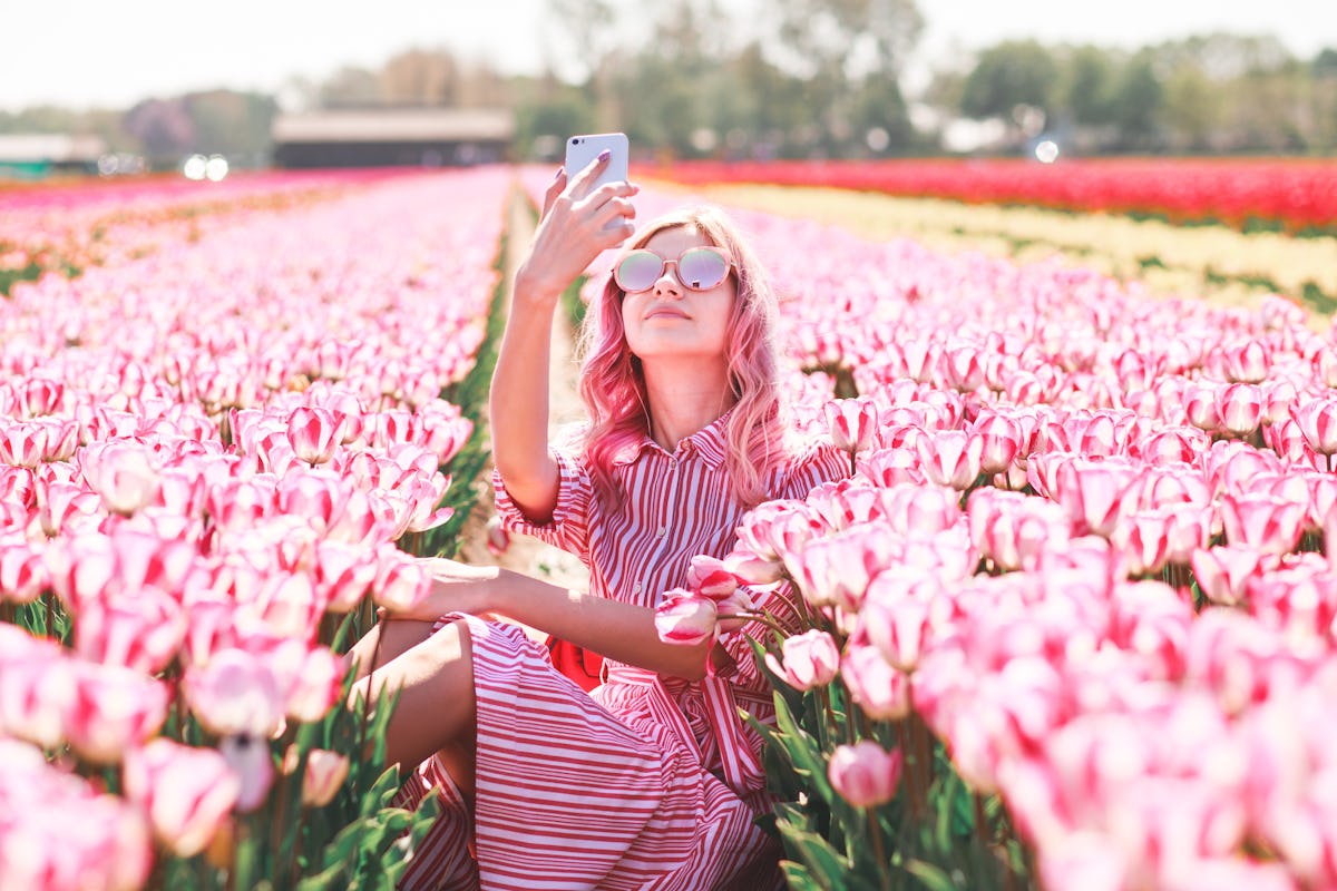 A woman in a pink striped dress and pink sunglasses takes a selfie in a field of pink tulips.