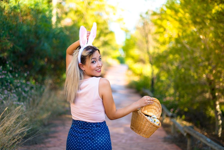 A woman walks down a trail with bunny ears on and holding a basket of Easter eggs.