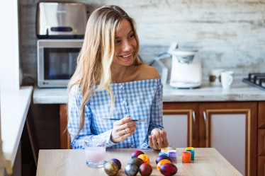 A woman with long blonde hair smiles while decorating Easter eggs in her kitchen.
