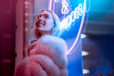 Happy smiling teen hipster girl in stylish glasses and fur looking up dreaming standing  near neon s...