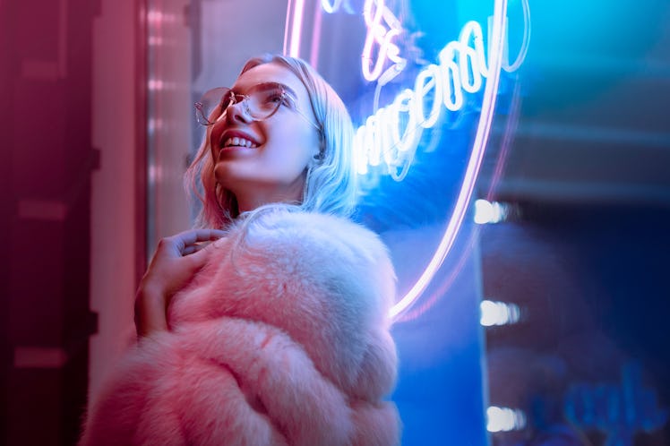 Happy smiling teen hipster girl in stylish glasses and fur looking up dreaming standing near neon s...