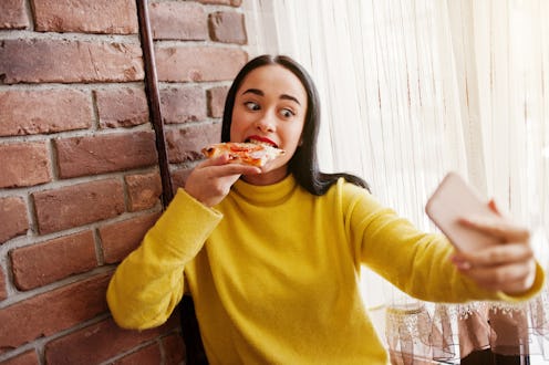 Funny brunette girl in yellow sweater eating pizza at restaurant and making selfie.
