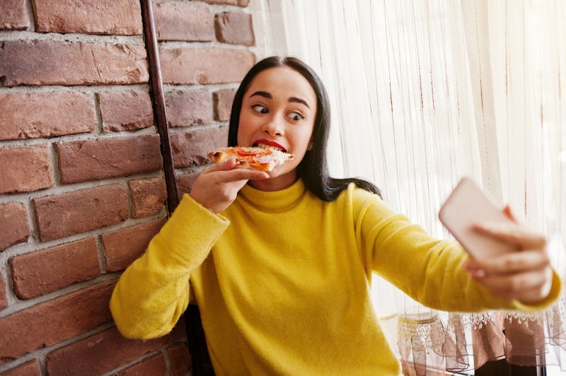 Funny brunette girl in yellow sweater eating pizza at restaurant and making selfie.