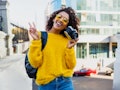 A happy woman, wearing a fuzzy yellow sweater and sunglasses throws up a peace sign while holding he...