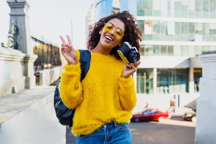 A happy woman, wearing a fuzzy yellow sweater and sunglasses throws up a peace sign while holding he...