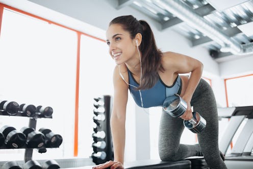 Young beautiful woman doing exercises with dumbbell in gym. Glad smiling girl is enjoying with her t...