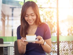 A happy woman holds a cup of coffee with a stroopwafel on top, while sitting in an outdoor cafe. 