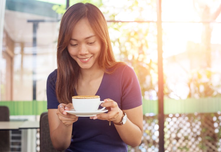 A happy woman holds a cup of coffee with a stroopwafel on top, while sitting in an outdoor cafe.