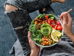 A woman in distressed gray jeans holds a colorful bowl of salad, veggies, and an avocado that's cut ...