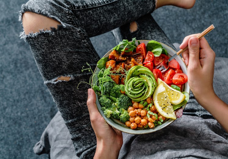 A woman in distressed gray jeans holds a colorful bowl of salad, veggies, and an avocado that's cut ...
