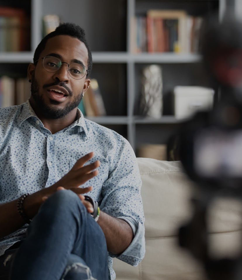 Millennial african hipster man blogger recording vlog on digital camera sit on sofa in living room, ...