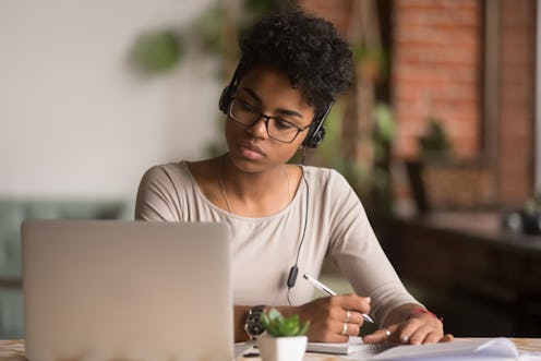 Focused mixed race woman wearing headphones watching webinar write notes study online with online te...