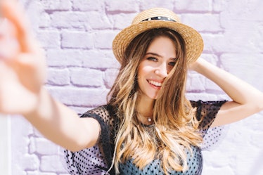 Close-up portrait of happy girl with mid-back length hair making selfie and holding straw hat. Outdo...