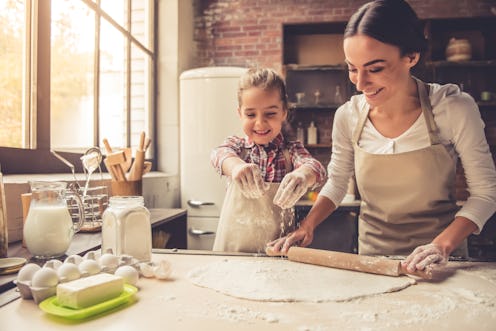 Cute little girl and her beautiful mother are flattening the dough using a rolling pin and smiling w...