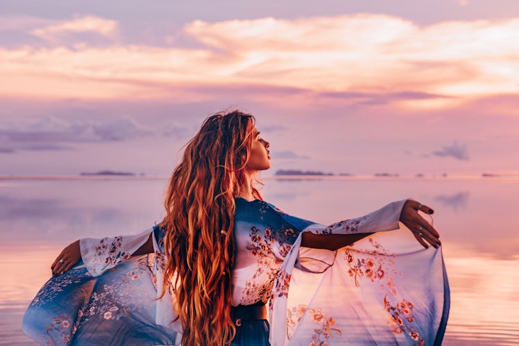 A woman in a floral, elegant dress walks on the beach at sunset.