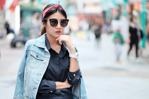 Asian girl wearing blue jean jacket, sun glasses and red headband walking on street with blurred bac...