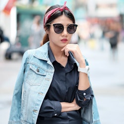 Asian girl wearing blue jean jacket, sun glasses and red headband walking on street with blurred bac...