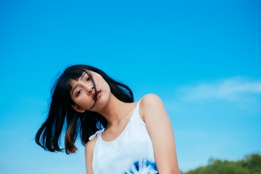 A young woman poses in a tie-dye tank top against a bright blue sky.
