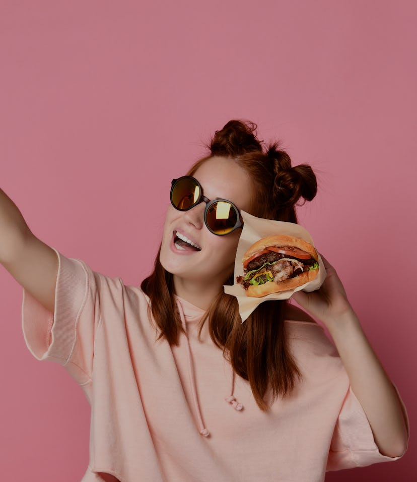 Positive ginger young woman pose for selfie, use modern cell phone, sit with burger in hand , have f...