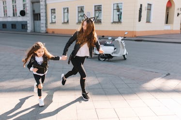 A young woman wearing black jeans, a leather jacket, and cat ears smiles and runs on a sidewalk whil...