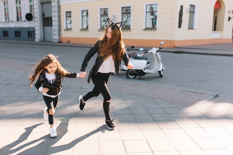 A young woman wearing black jeans, a leather jacket, and cat ears smiles and runs on a sidewalk whil...
