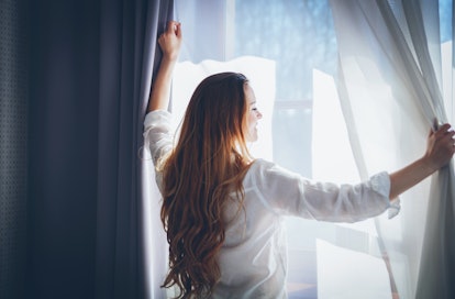 Pretty young woman in modern apartment opening window curtains after wake up