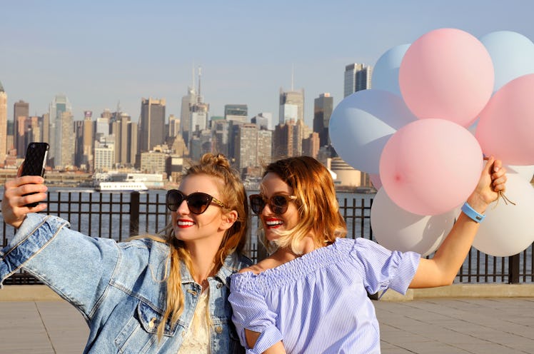 Two happy friends take a selfie, while holding balloons with the New York City skyline in the backgr...