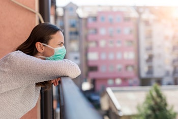 Portrait of young woman in home isolation standing on a balcony, wearing surgical mask outdoors as p...