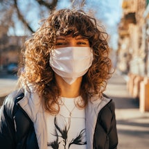 Happy young woman wearing facial mask for virus protection standing outdoors on sunny spring day.