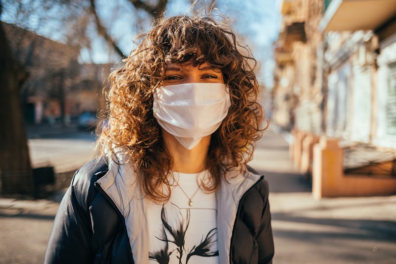 Happy young woman wearing facial mask for virus protection standing outdoors on sunny spring day.