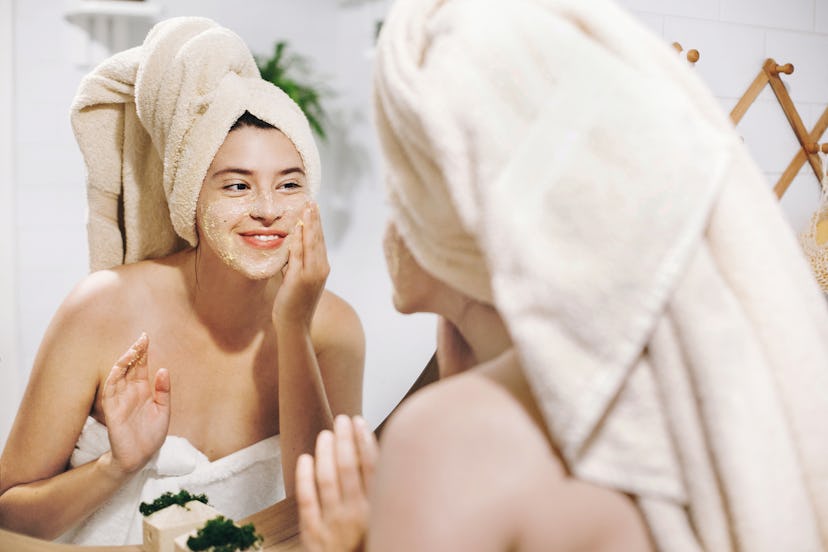 A woman smiling and applying a face mask while looking at mirror with her hair wrapped in a towel as...