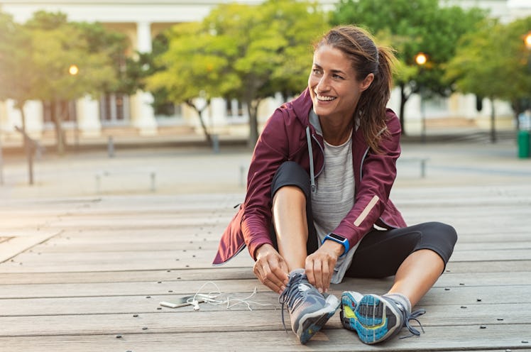 Mature fitness woman tie shoelaces on road. Cheerful runner sitting on floor on city streets with mo...
