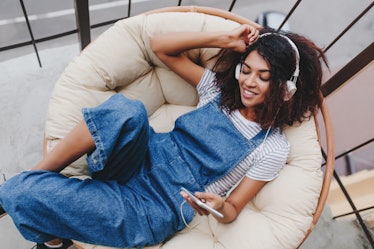 Blissful black young woman in vintage denim clothes chilling on terrace in white headphones. Attract...
