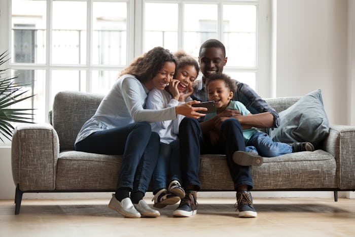 Happy african family with 2 children son daughter having fun with gadget on couch using smartphone a...