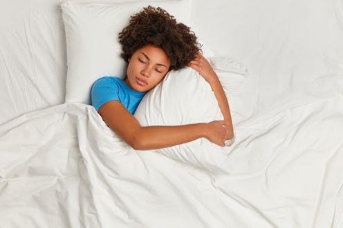 Dark skinned restful woman lying in bed and sleeps peacefully, hugs pillow, enjoys comfort and softn...