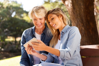 A blonde woman shows something on her cell phone to her mom while they sit on a bench in the sun.