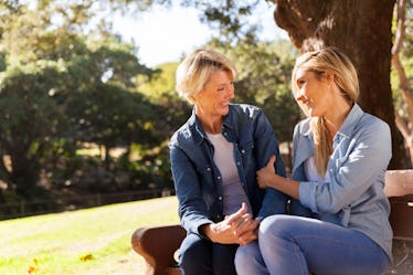 A blonde mother and her daughter smile and chat on a bench in the sunshine.