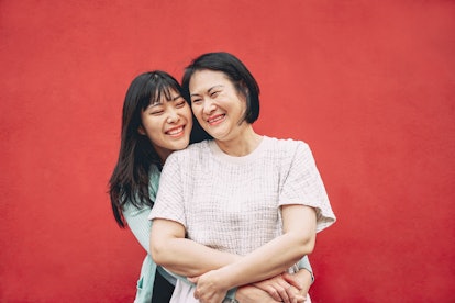 A happy asian mother and daughter hug in front of a red wall.