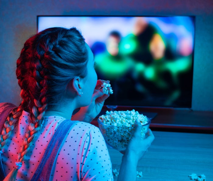 A young girl watching movies and eating popcorn with a bowl on the background of the TV. The color b...