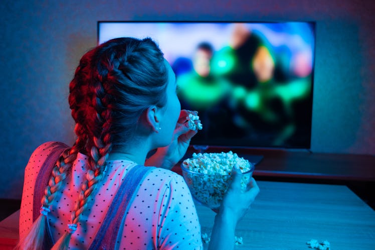 A young girl watching movies and eating popcorn with a bowl on the background of the TV. The color b...
